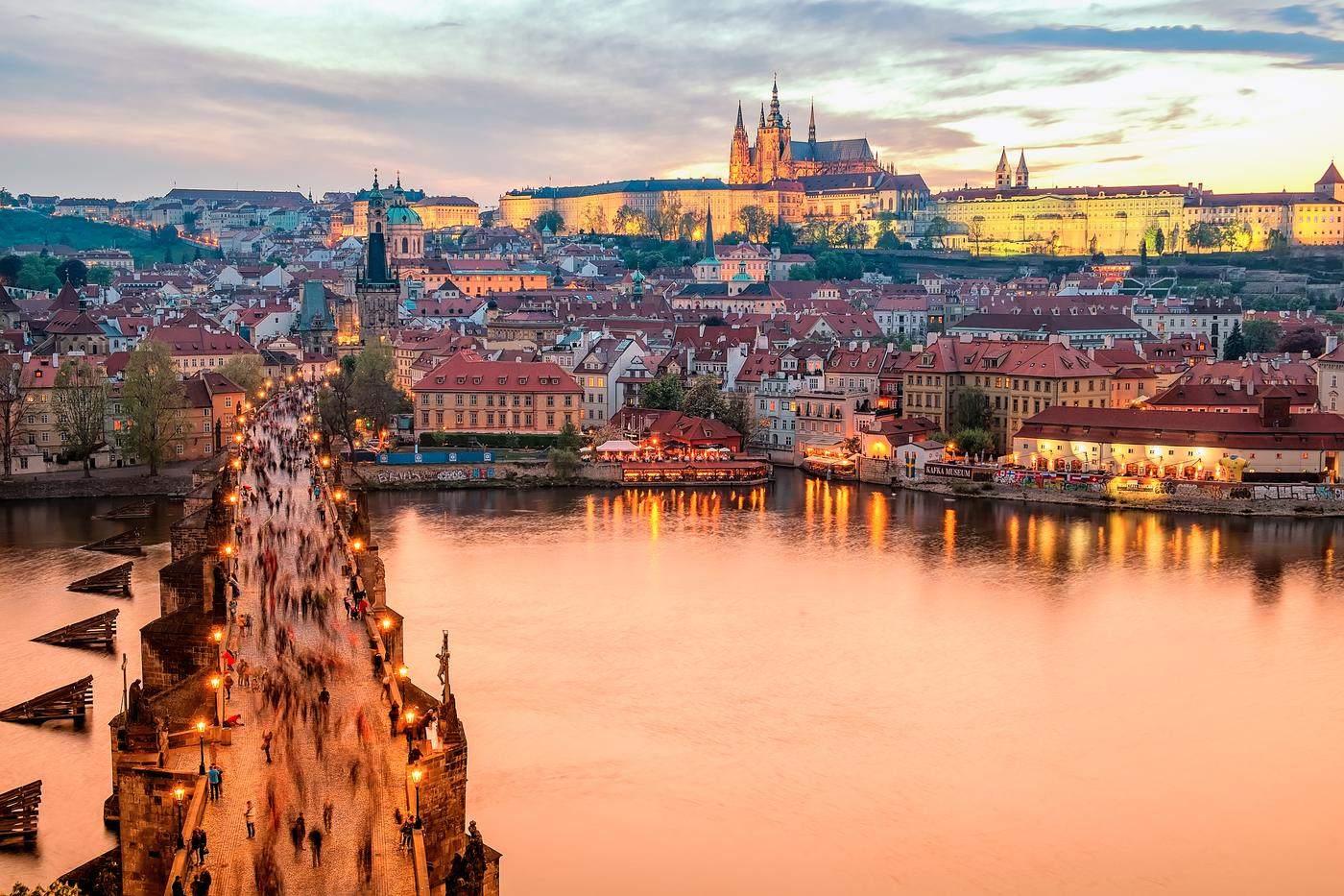 Crowds crossing a lamplit stone bridge toward a hilltop castle at dusk
