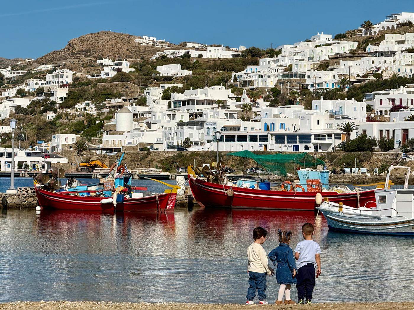 Three small children watching red fishing boats in a whitewashed harbor