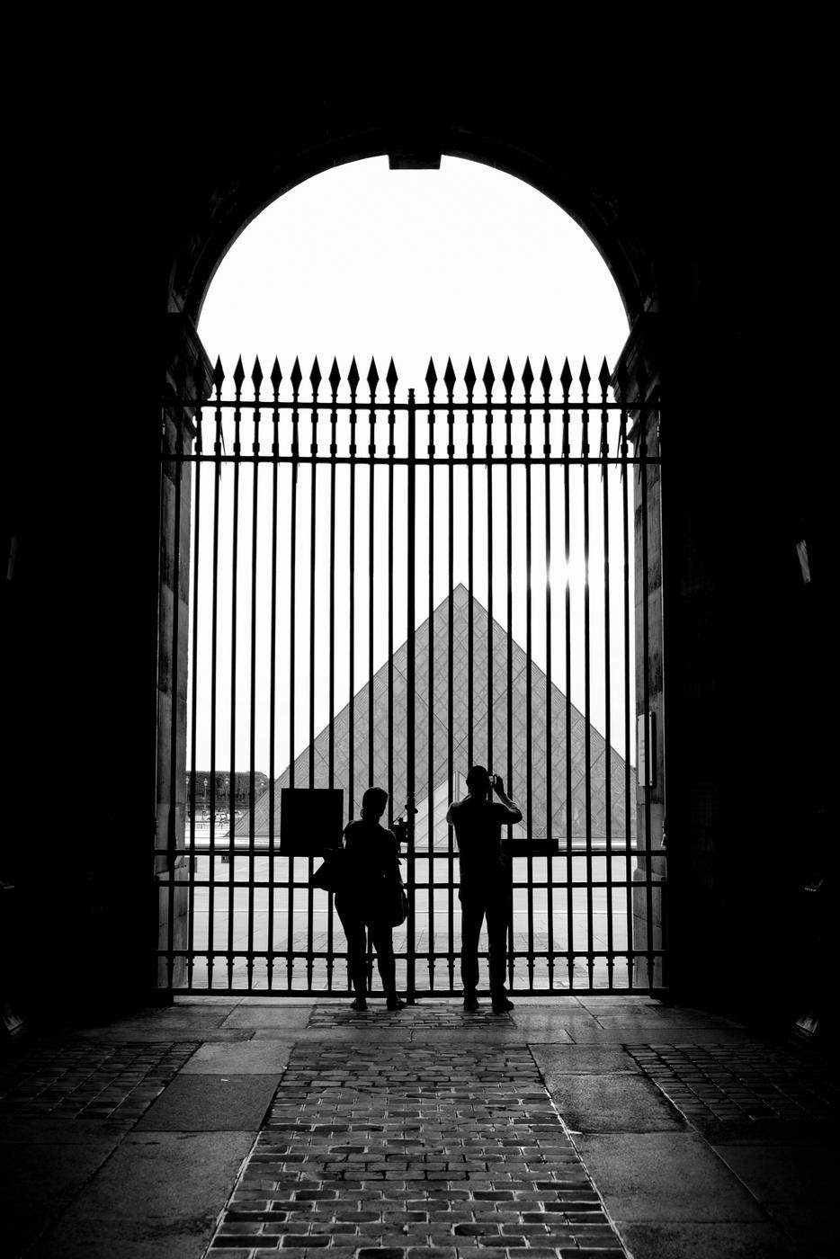 Two figures at an iron gate beneath an arch, a glass pyramid beyond